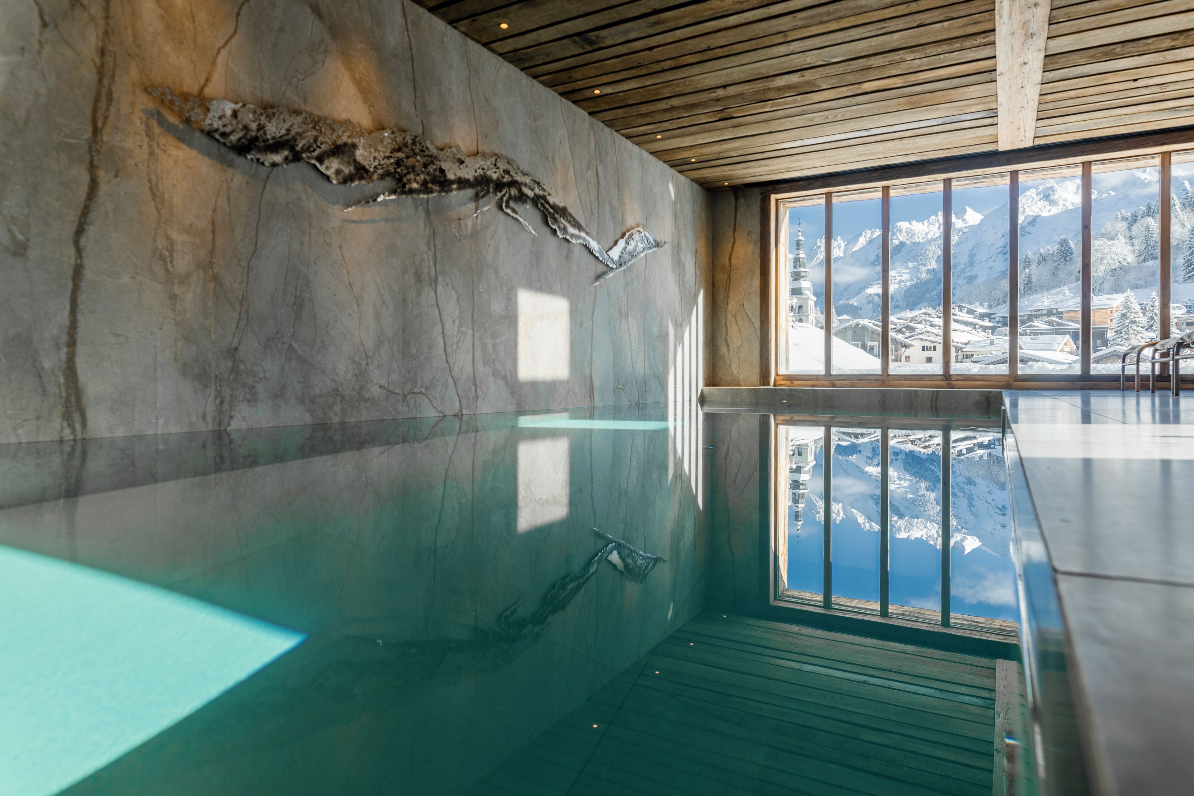 An indoor swimming pool with a wooden ceiling and a view of snow-covered mountains through large windows.