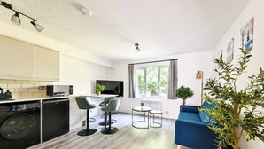A modern kitchen with a dishwasher, a dining area with two chairs, a small table, and a blue sofa. There is a window with curtains and a plant on the right. - Charming 1-Bed Flat in London – 3 Mins to Tube + Free Parking (London)
