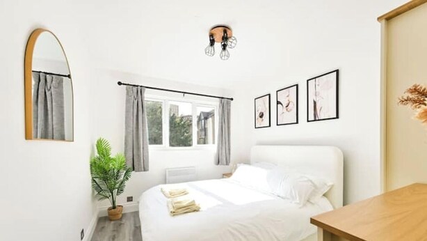 A bedroom with a bed, a potted plant, a window with curtains, and framed pictures on the wall. - Charming 1-Bed Flat in London – 3 Mins to Tube + Free Parking (London)