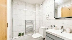 A modern bathroom with white marble tiles, a glass shower enclosure, a toilet, and a vanity with a mirror. - Charming 1-Bed Flat in London – 3 Mins to Tube + Free Parking (London)