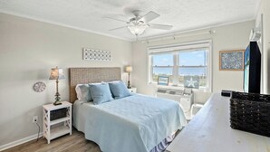 A bedroom with a bed, nightstands, a ceiling fan, a window with a view, and a desk with a chair. - Palmwood Villas 101- Oceanfront 2BR/2BA- Pool (North Myrtle Beach)