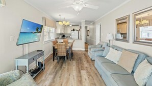 A modern living room with a TV, a dining area, and a kitchen in the background. - Palmwood Villas 101- Oceanfront 2BR/2BA- Pool (North Myrtle Beach)