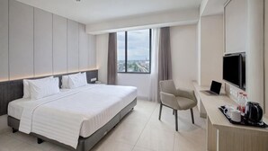 Ein Hotelzimmer mit einem Bett, einem Schreibtisch mit Computer, einem Stuhl, einem Fernseher und einem Fenster mit Blick auf die Stadt.