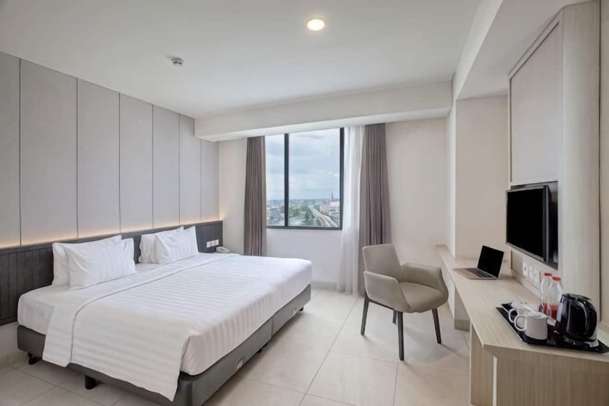 Ein Hotelzimmer mit einem Bett, einem Schreibtisch mit Computer, einem Stuhl, einem Fernseher und einem Fenster mit Blick auf die Stadt.