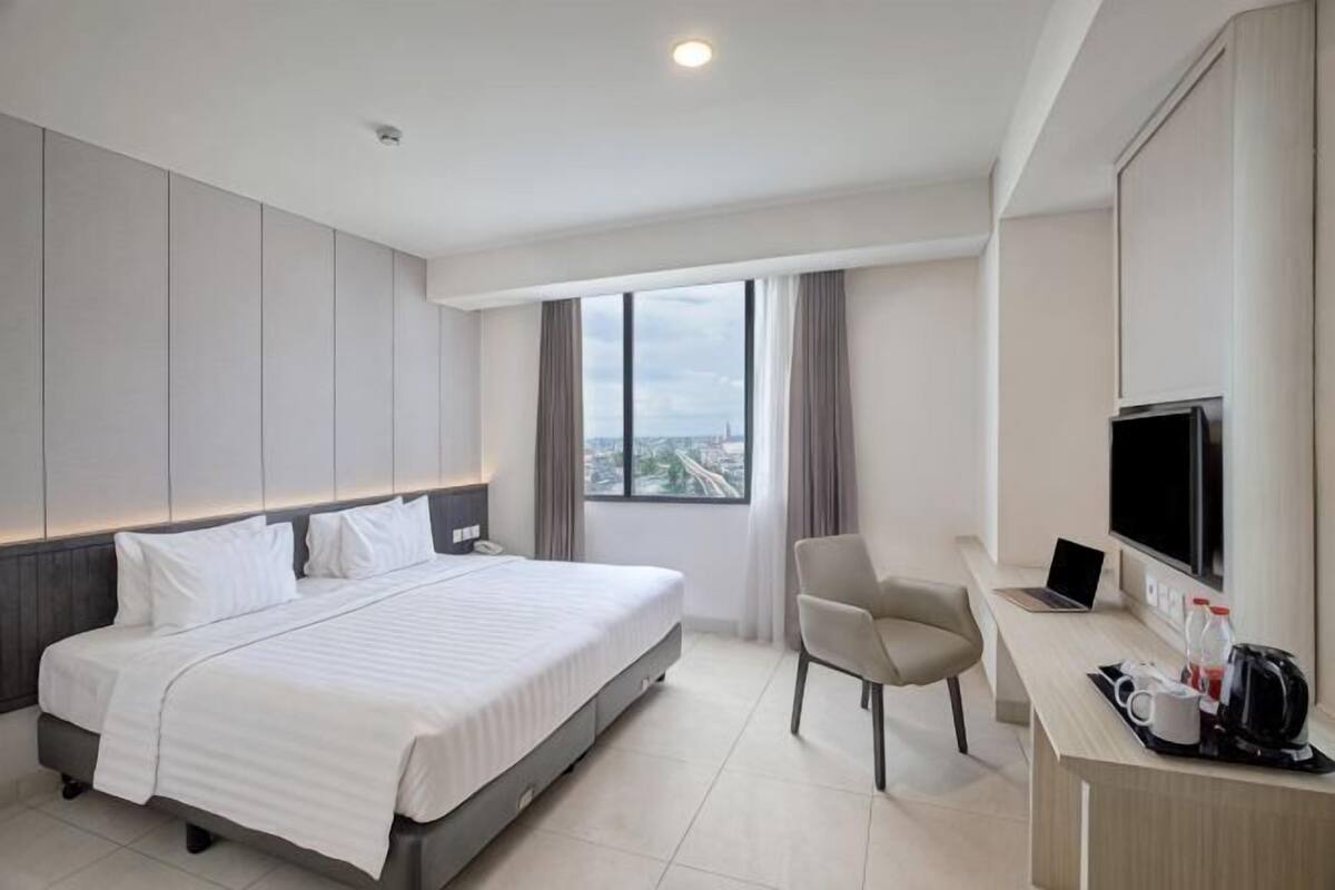 A hotel room with a bed, a desk with a computer, a chair, a TV, and a window with a city view.
