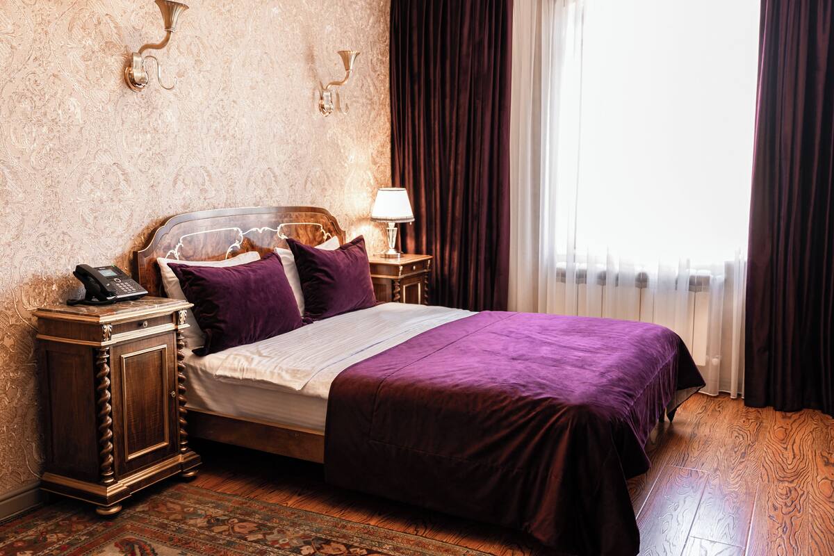 Un lit bien fait, recouvert d’une couvre-lit violette, une table de chevet en bois avec une lampe et un téléphone.
