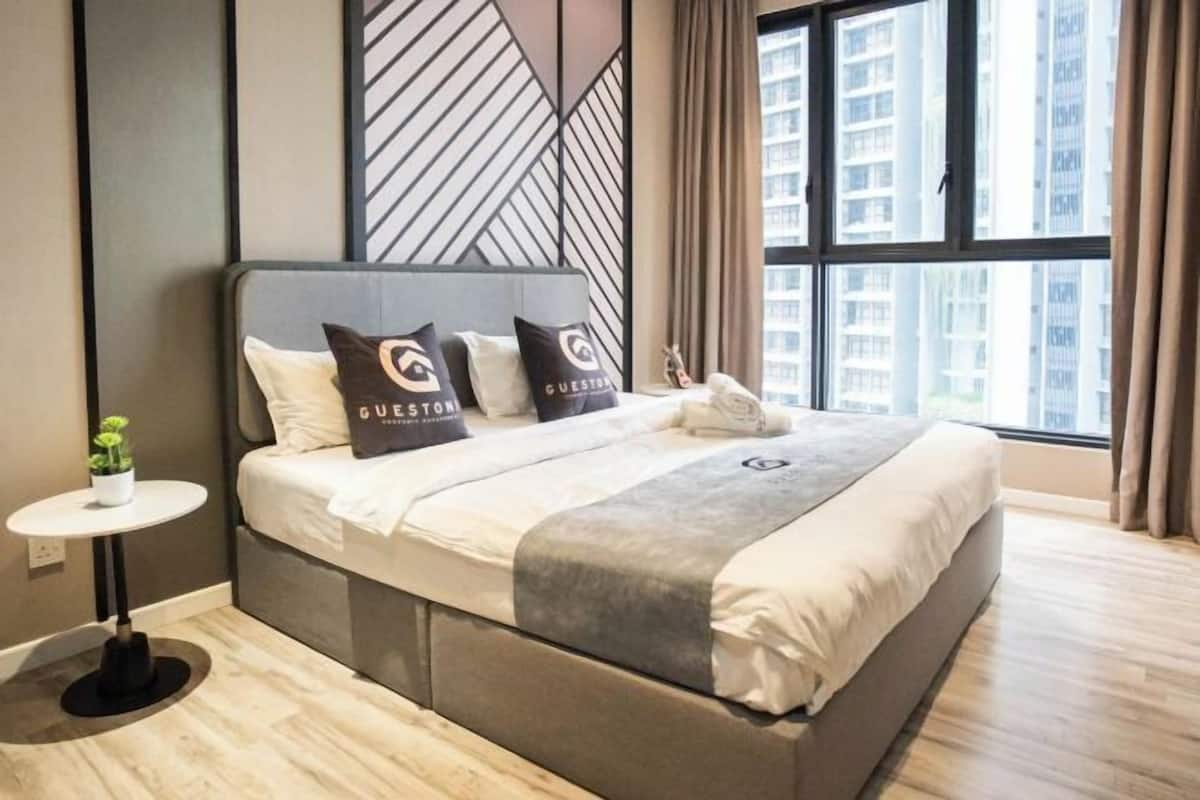 A modern hotel room with a large bed, a small round table with a plant, and a view of tall buildings through large windows.