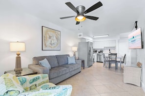 A living room with a sofa, a coffee table, a ceiling fan, a TV, and a kitchen in the background.