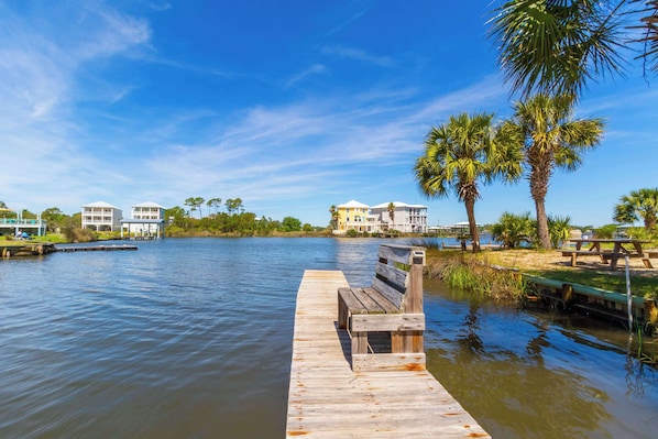A wooden dock extending into calm water with a bench, surrounded by palm trees and buildings in the background.