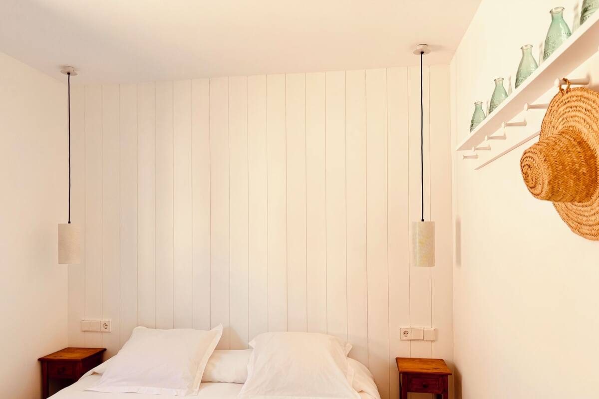 A bedroom with a bed, two bedside tables, a wall-mounted shelf with bottles, and two pendant lights.