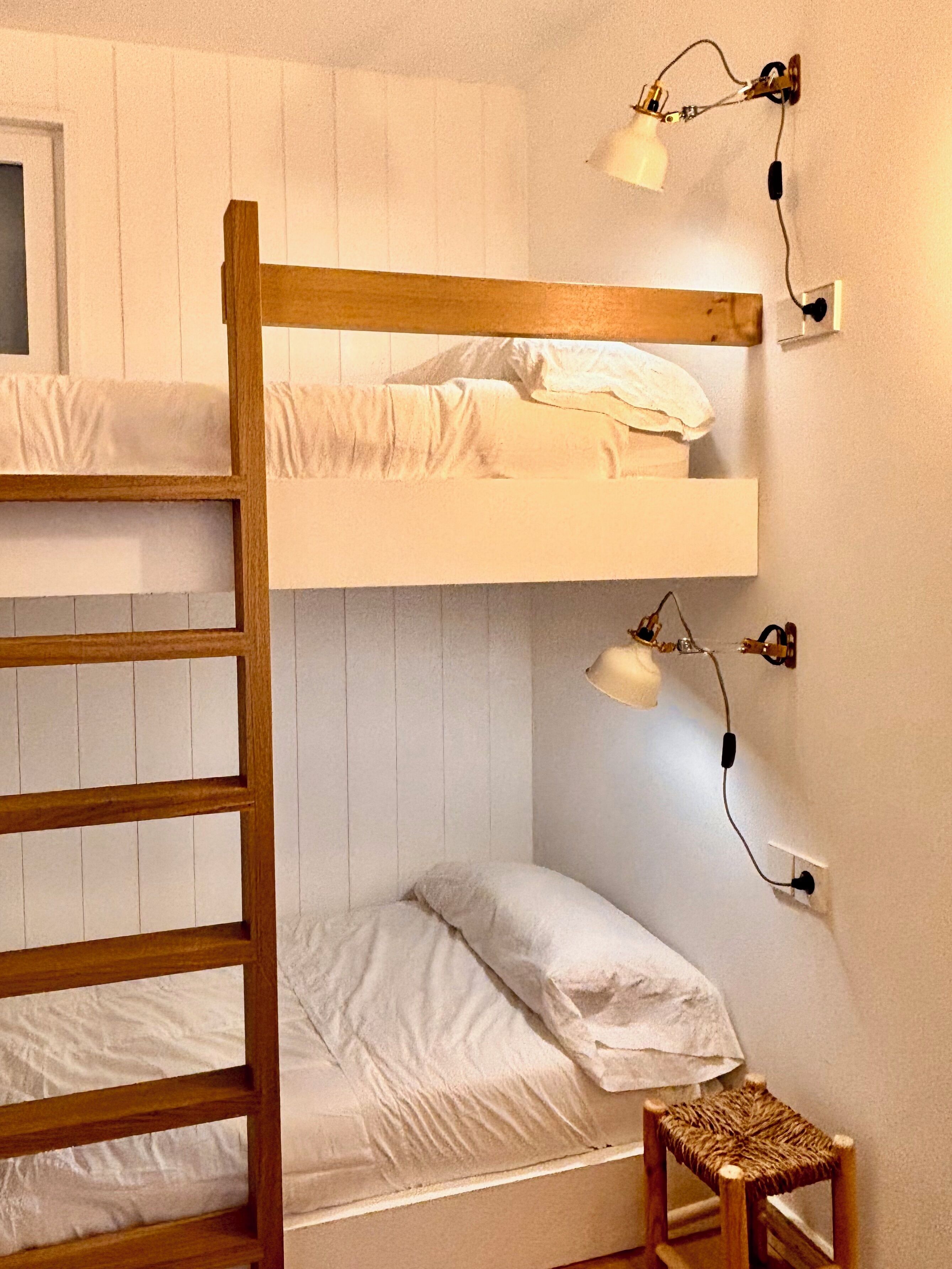 Una cama litera con escalera, un taburete pequeño y lámparas fijadas a la pared.