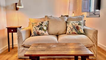 Un sofá con cojines estampados, una mesa de centro de madera, una mesita auxiliar y una lámpara de pared, junto con un televisor.