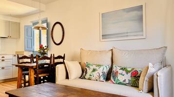 A living room with a wooden table, a sofa with floral cushions, a painting on the wall, and a kitchen in the background.