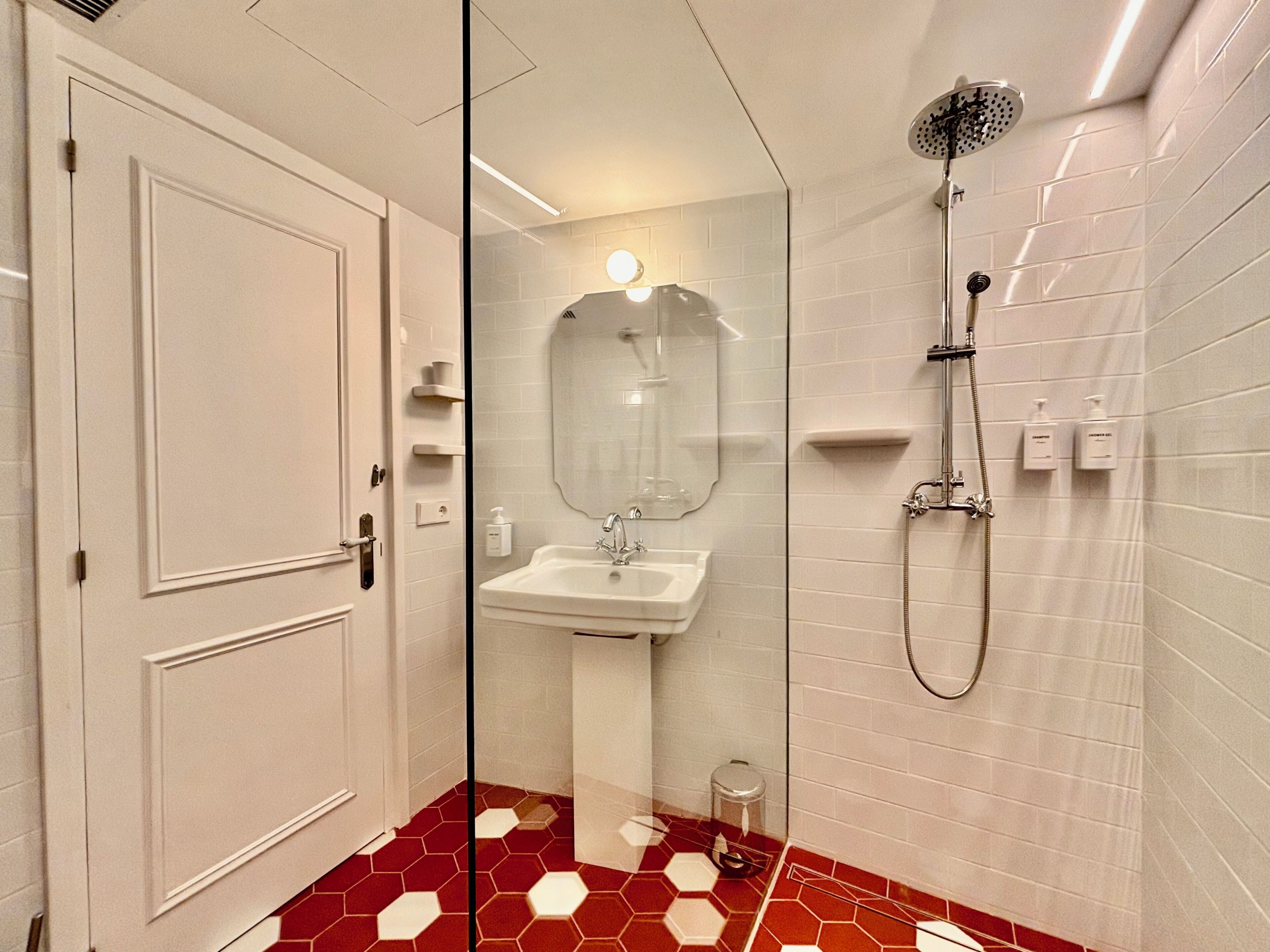 A bathroom with a white door, a sink, a mirror, a shower, and a red and white tiled floor.