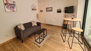 Ett modernt vardagsrum med en soffa, ett soffbord och barstolar. En tv är monterad på väggen, och det finns inramade tavlor och en affisch på väggen.