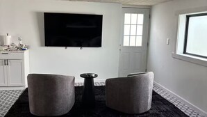 Ein modernes Wohnzimmer mit einem schwarzen Teppich, zwei grauen Hocker, einem kleinen schwarzen Tisch und einem an der Wand befestigten Fernseher.