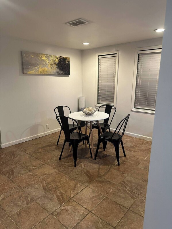 A dining area with a round table and four chairs, a painting on the wall, and two windows with blinds. - Private house 1 bedroom 1 bath ground-floor (Warren)