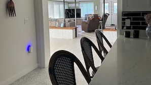 Eine moderne Küche mit Deckenventilator in Weiß, einem Essbereich mit Marmorplatte und Korbstühlen sowie Blick in ein Wohnzimmer mit Sofa und Fernseher.