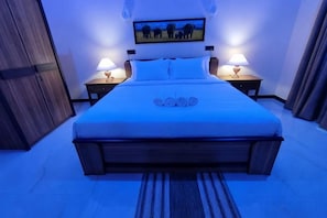 A bed with a headboard, two bedside tables with lamps, a wall-mounted picture, and a wooden floor.