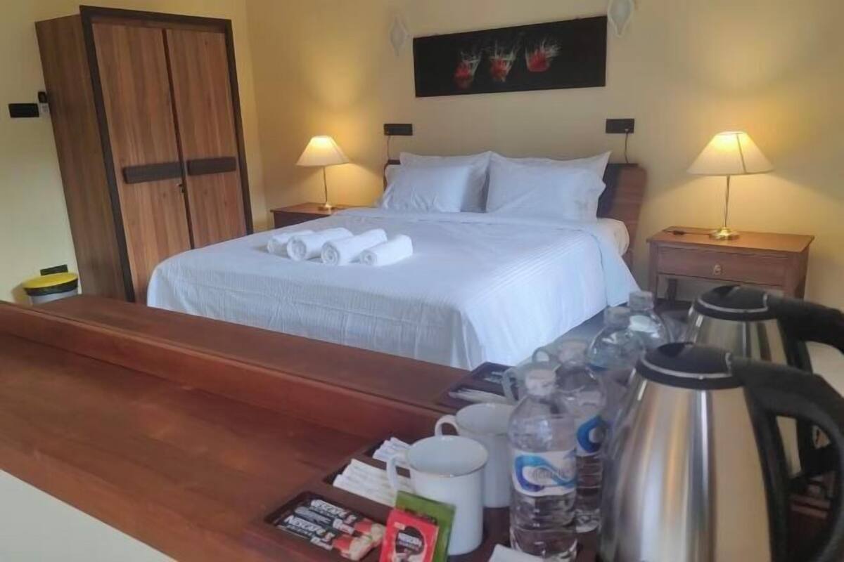 A neatly made bed with rolled towels, a wooden desk with a coffee pot, mugs, and a water bottle.