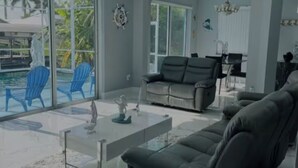A modern living room with a large glass door leading to a pool area, a black leather sofa, a glass coffee table, and a dining area with a chandelier. - 4-bedroom, pool, fishing, games, BBQ, kayak and front water. (Cape Coral)