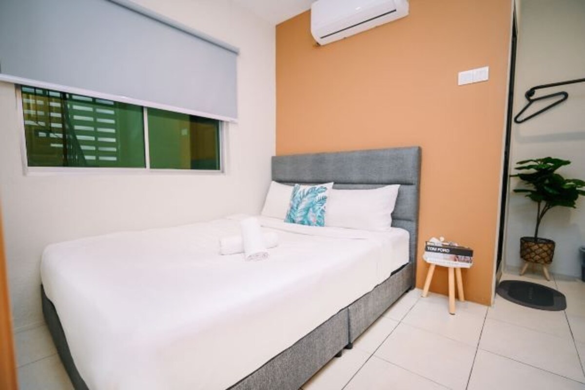 A bedroom with a bed, a window with blinds, an air conditioning unit, a small table with a magazine, and a potted plant.