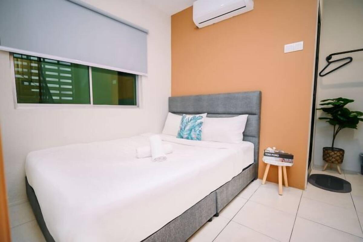 A bedroom with a bed, a window with blinds, an air conditioning unit, a small table with a magazine, and a potted plant.