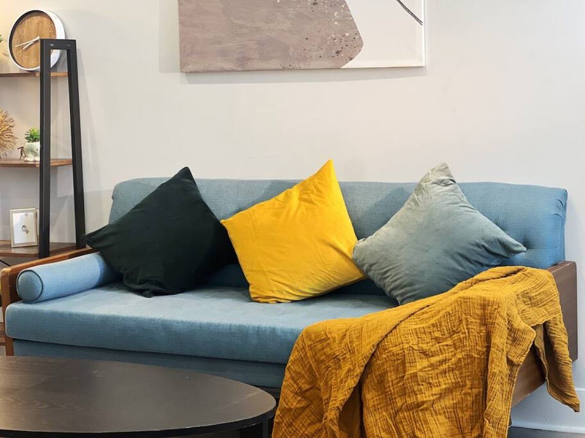 A blue sofa with three pillows in black, yellow, and grey, a mustard-colored throw blanket, and a black coffee table.