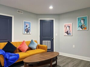 A living room with a yellow sofa, a dark door, and two framed pictures on the wall. - Ramblei near SJU & CHOP | 8-Bedroom House (Philadelphia)