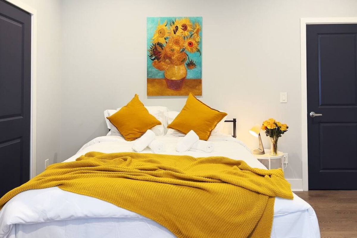 A bed with white bedding and two mustard pillows, a yellow throw blanket, a painting of sunflowers on the wall, and a small table with a lamp and flowers.