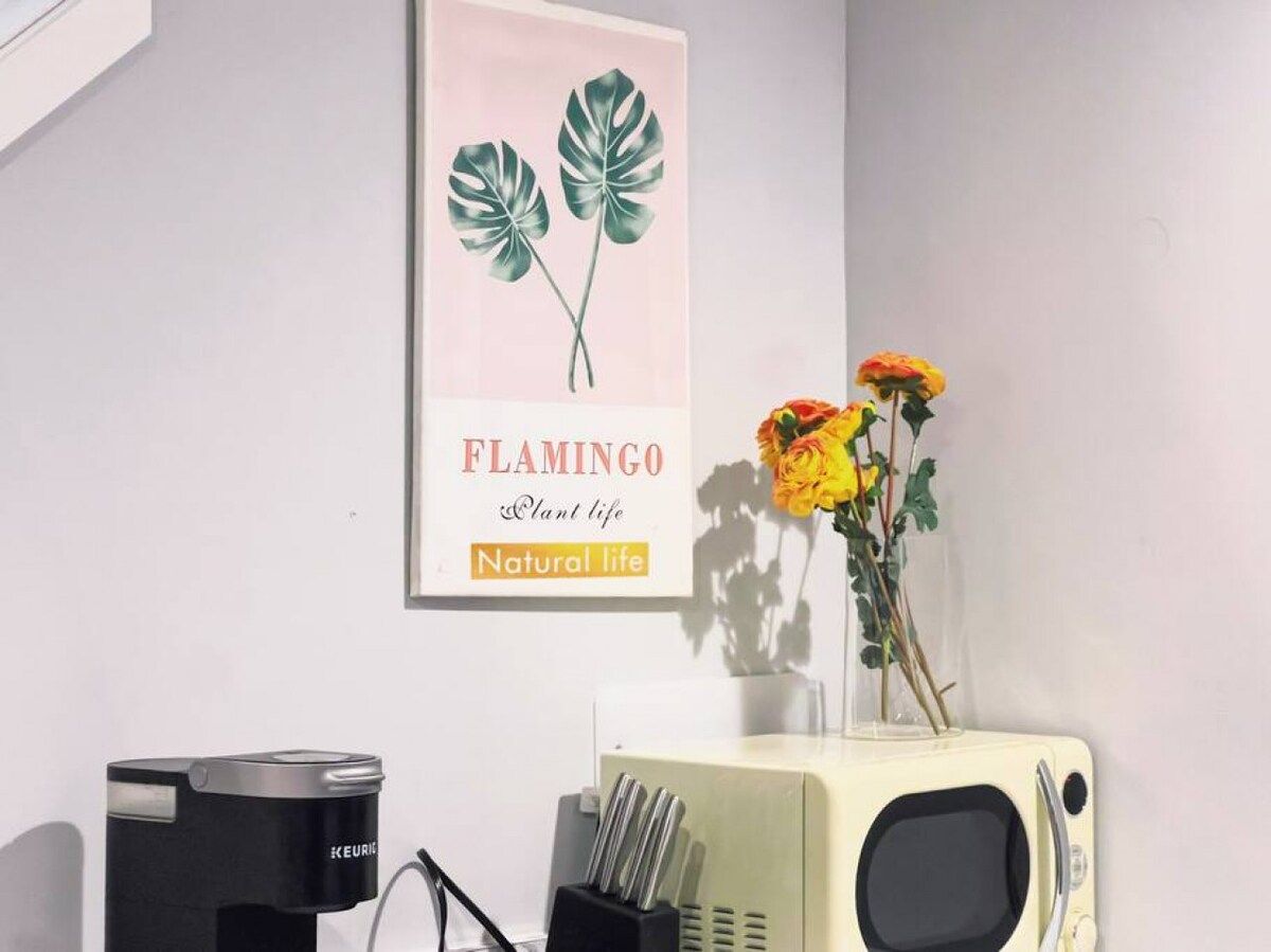 Una habitación con una pared blanca, un cartel enmarcado de una planta, una radio antigua y un florero con flores.