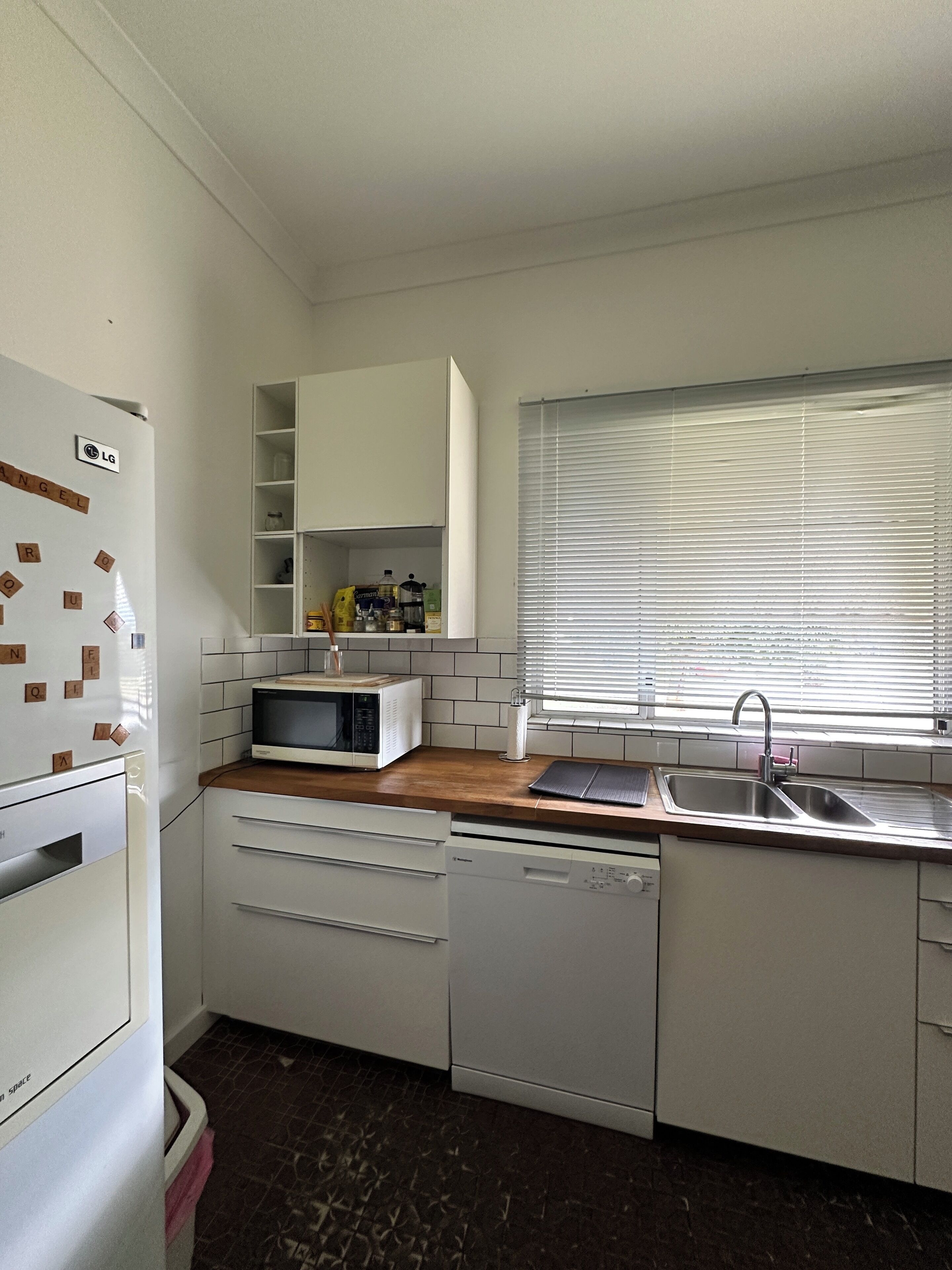 Una cocina compacta con gabinetes blancos, microondas, lavavajillas y ventana con persianas.
