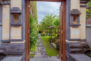 A pathway leading to a traditional structure with a wooden door, surrounded by greenery and a clear sky. - Puri Canggu Rooms 2 (Bali)