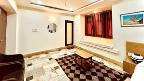 Una habitación con piso a cuadros, un espejo redondo, una ventana con cortinas rojas, una cama, una mesita y un cuadro en la pared.
