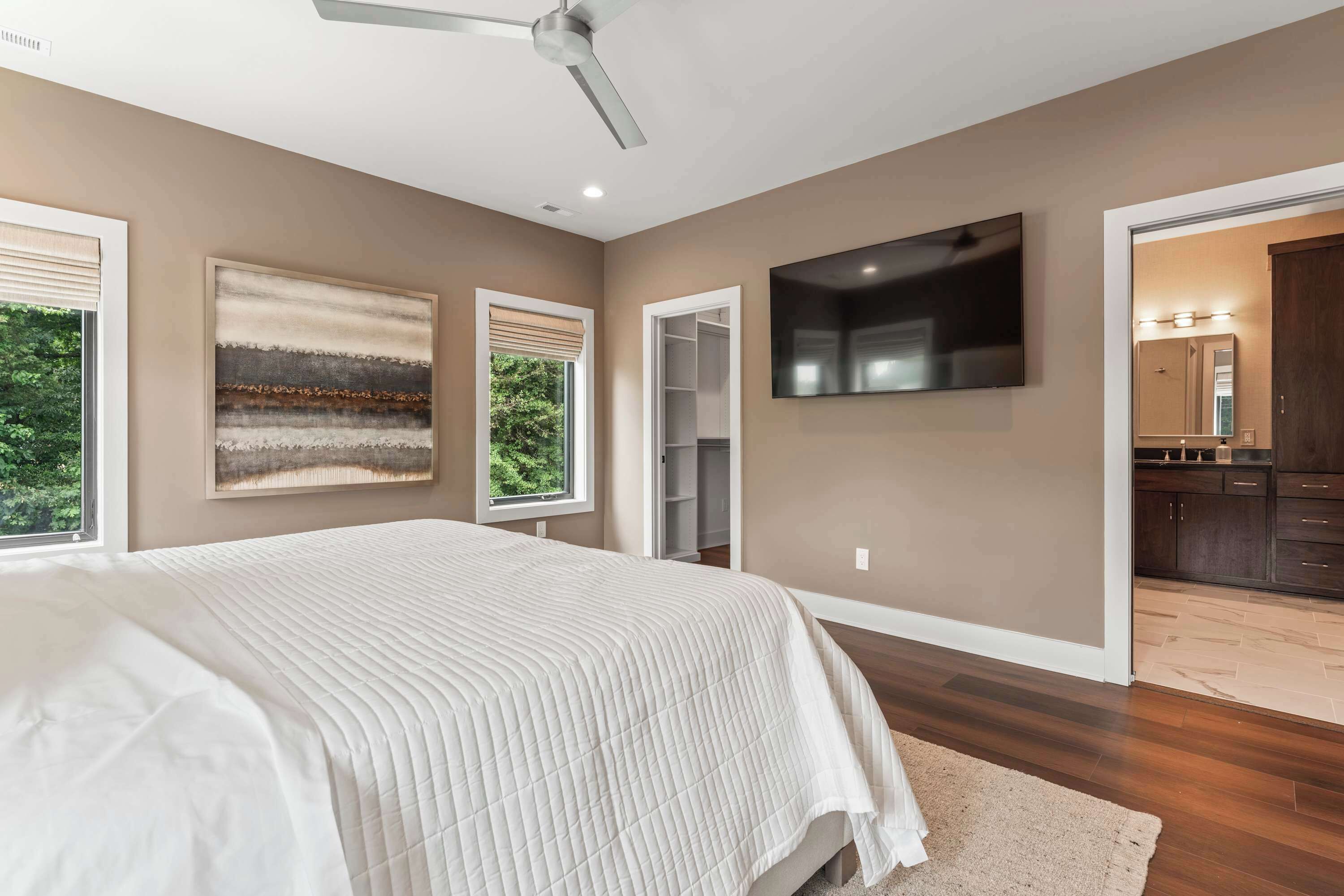 Une chambre avec un grand lit, un ventilateur de plafond, un téléviseur fixé au mur et une vue sur la cuisine par une porte ouverte.