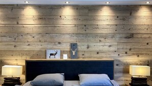 A bedroom with a wooden headboard, two bedside tables with lamps, a bed with two pillows, and a wall-mounted shelf with a deer picture and a wooden plaque. - Spacious 3.5-Room | Best Location • Free Parking (Leukerbad)
