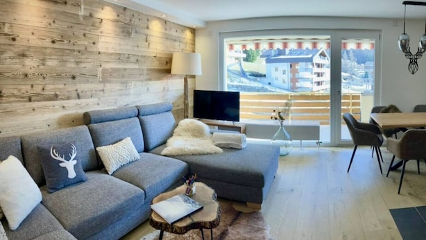 A modern living room with a grey sectional sofa, a flat-screen TV, a dining area with a wooden table and chairs, and a large window with a view of buildings and trees. - Spacious 3.5-Room | Best Location • Free Parking (Leukerbad)