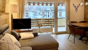 A modern living room with a flat-screen TV, a sofa, a dining table with chairs, and a balcony with a view of buildings. - Spacious 3.5-Room | Best Location • Free Parking (Leukerbad)