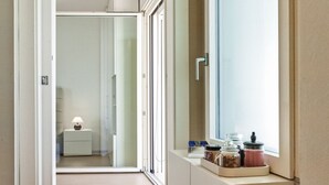 Un couloir avec une porte-fenêtre coulissante donnant sur une salle de bain, une petite table de chevet avec une lampe et une fenêtre sur le rebord de laquelle sont posés des pots.