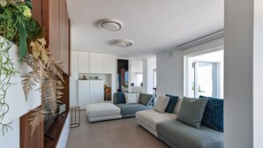 Un salon moderne avec un canapé gris, une petite table basse et un meuble en bois. On aperçoit à l’arrière-plan un coin cuisine avec des meubles blancs et une fenêtre.