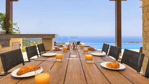 Mesa de madera dispuesta para el desayuno, con sillas, platos, vasos y croissants, con vistas a un paisaje marino.
