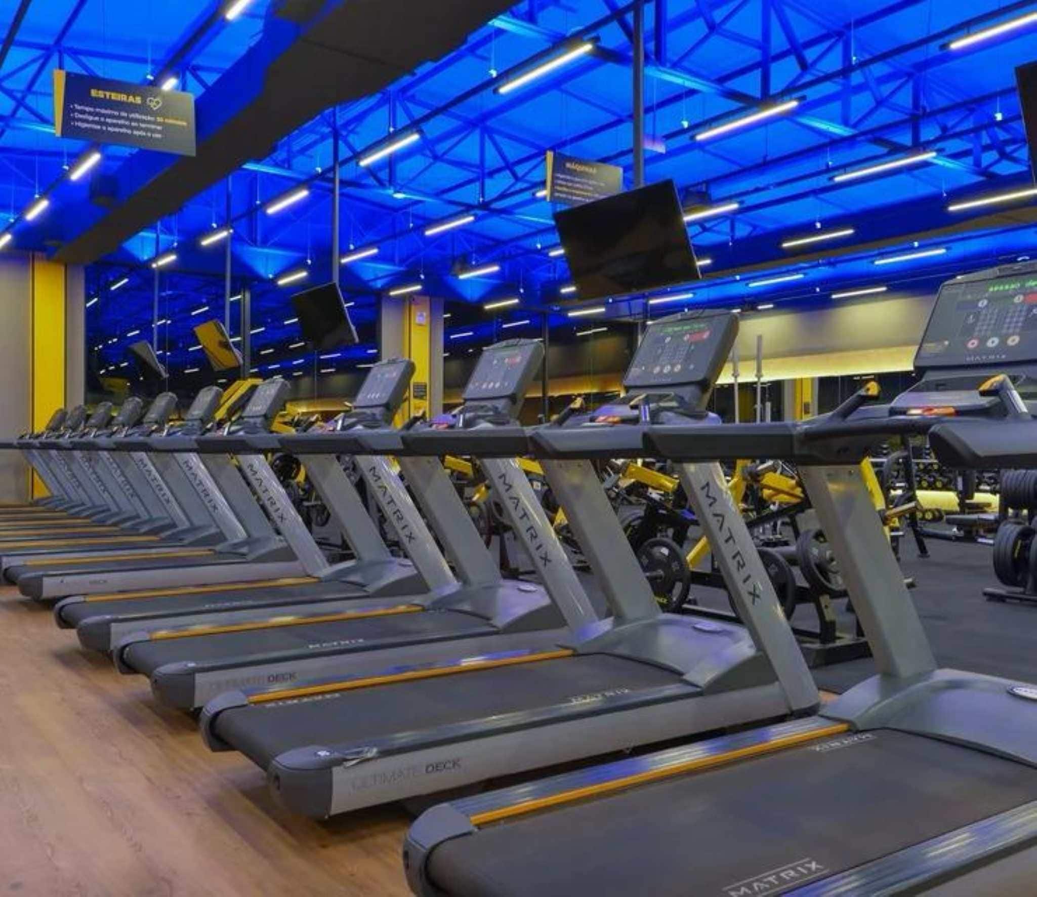 Varias caminadoras con pantallas digitales, bajo luces azules en un gimnasio.