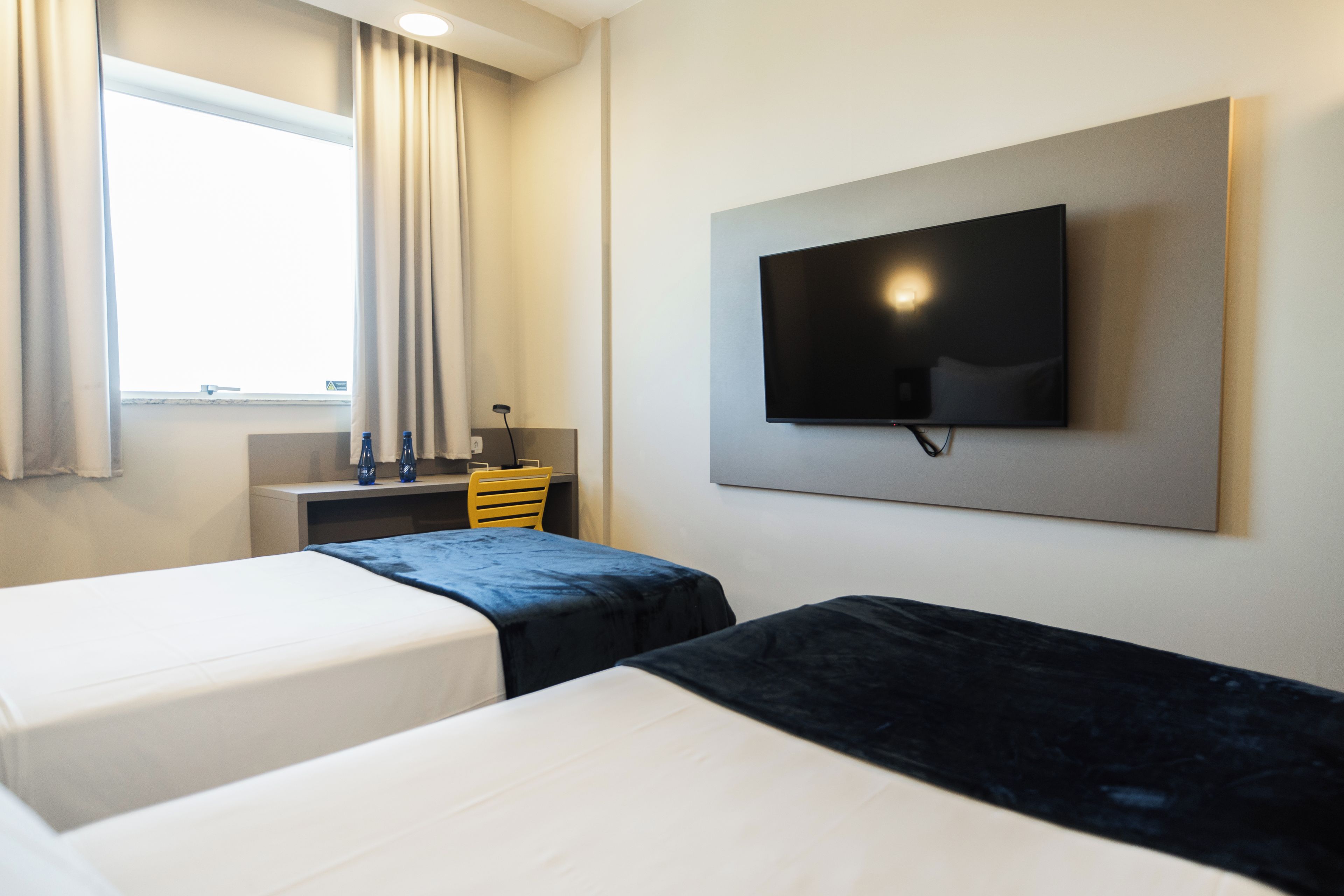 Habitación de hotel con dos camas, un televisor de pantalla plana montado en la pared, un escritorio pequeño con una silla y una ventana con cortinas.