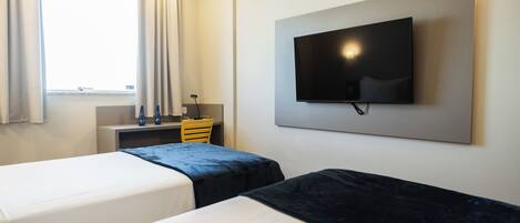 Habitación de hotel con dos camas, un televisor de pantalla plana montado en la pared, un escritorio pequeño con una silla y una ventana con cortinas.