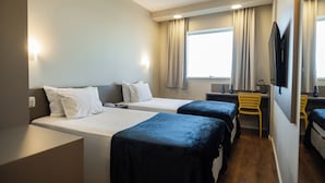Une chambre d’hôtel avec deux lits simples, un bureau, une chaise, une télévision et une fenêtre avec des rideaux.