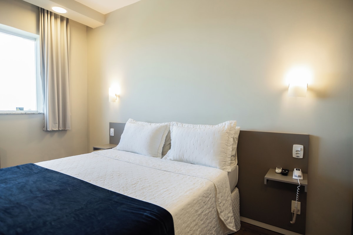 A hotel room with a bed, a bedside table, a nightstand, a wall-mounted phone, and a window with curtains.