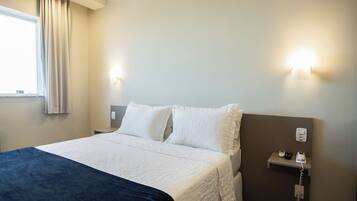 A hotel room with a bed, a bedside table, a nightstand, a wall-mounted phone, and a window with curtains.