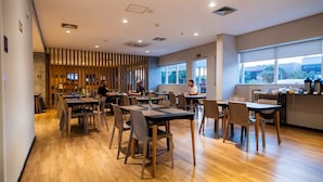 Un espace repas moderne avec un plancher en bois, des tables et des chaises. De grandes fenêtres laissent entrer la lumière naturelle et offrent une vue sur l’extérieur.