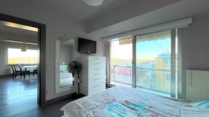 Une chambre avec un grand lit, un téléviseur fixé au mur, une fenêtre donnant sur des immeubles et une chaise près d’une table.