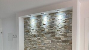 A modern living room with a stone wall, a flat-screen TV, and a white entertainment unit. - New Studio | Dream Views • Balcony • Free Parking (Leukerbad)